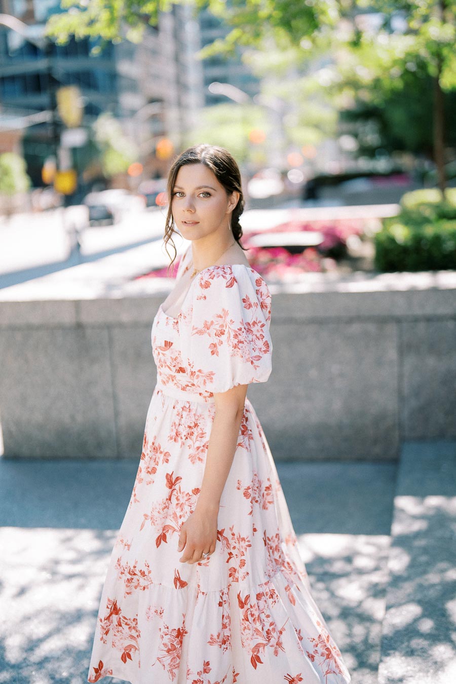 A woman in a floral dress standing outside on a sunny day in a city setting.