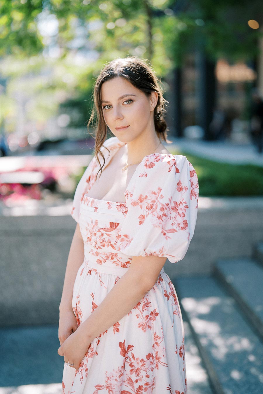 Young woman in a floral dress standing outdoors with a blurred green and urban background, exuding elegance and serenity.