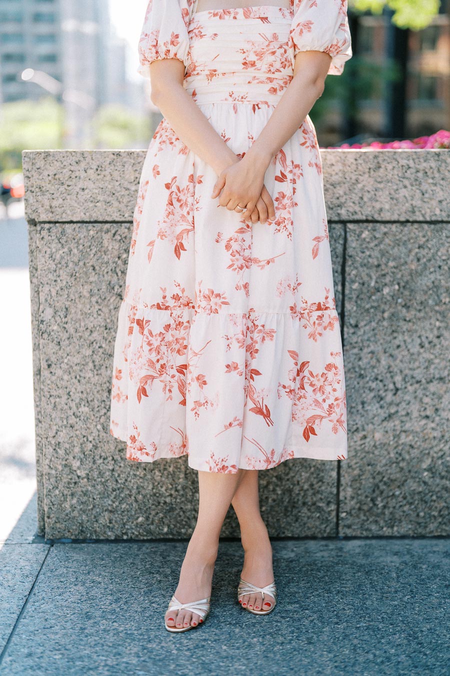 Woman in a floral summer dress and sandals standing in an urban setting.