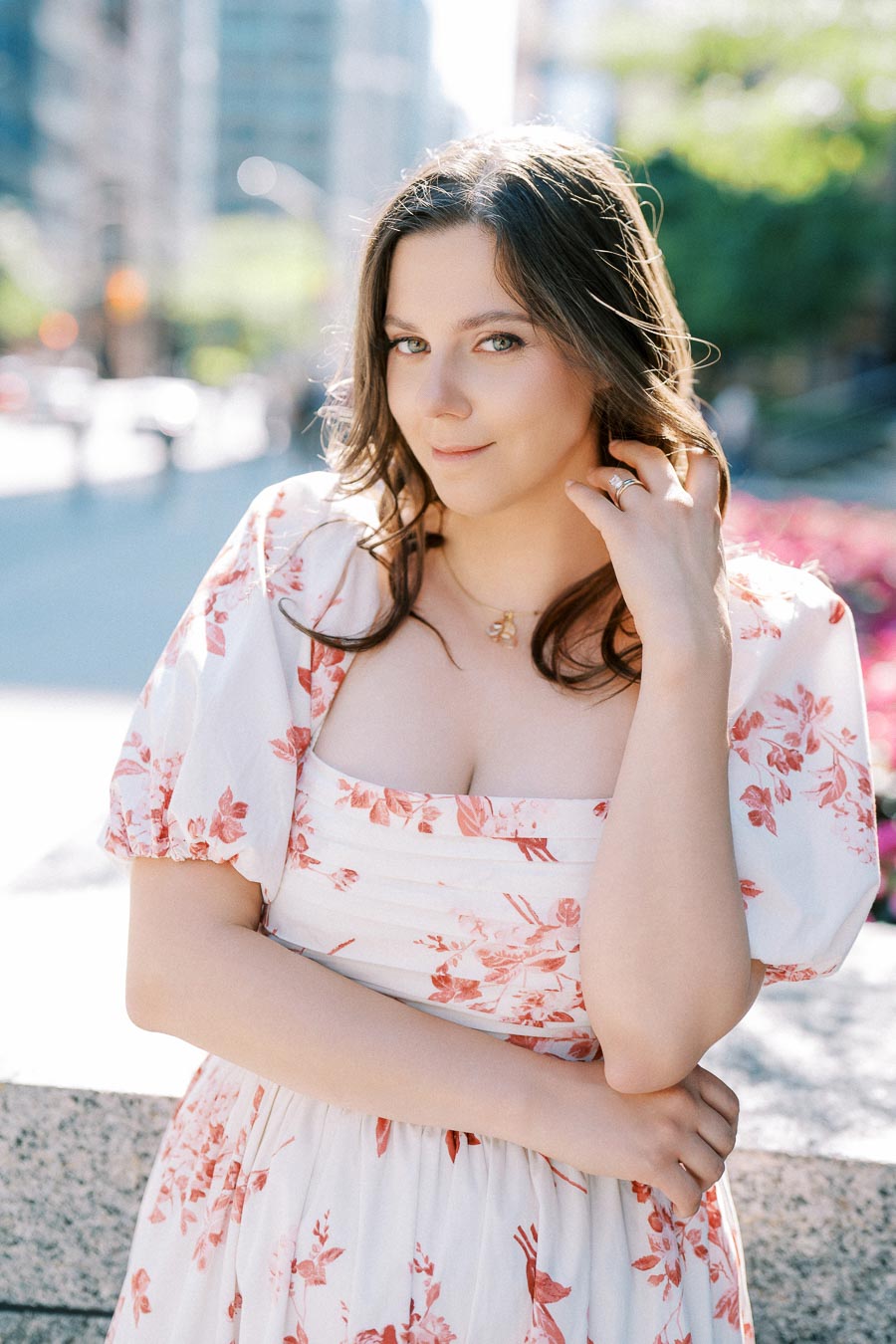 Young woman in a floral dress posing outdoors in a sunlit urban park setting.