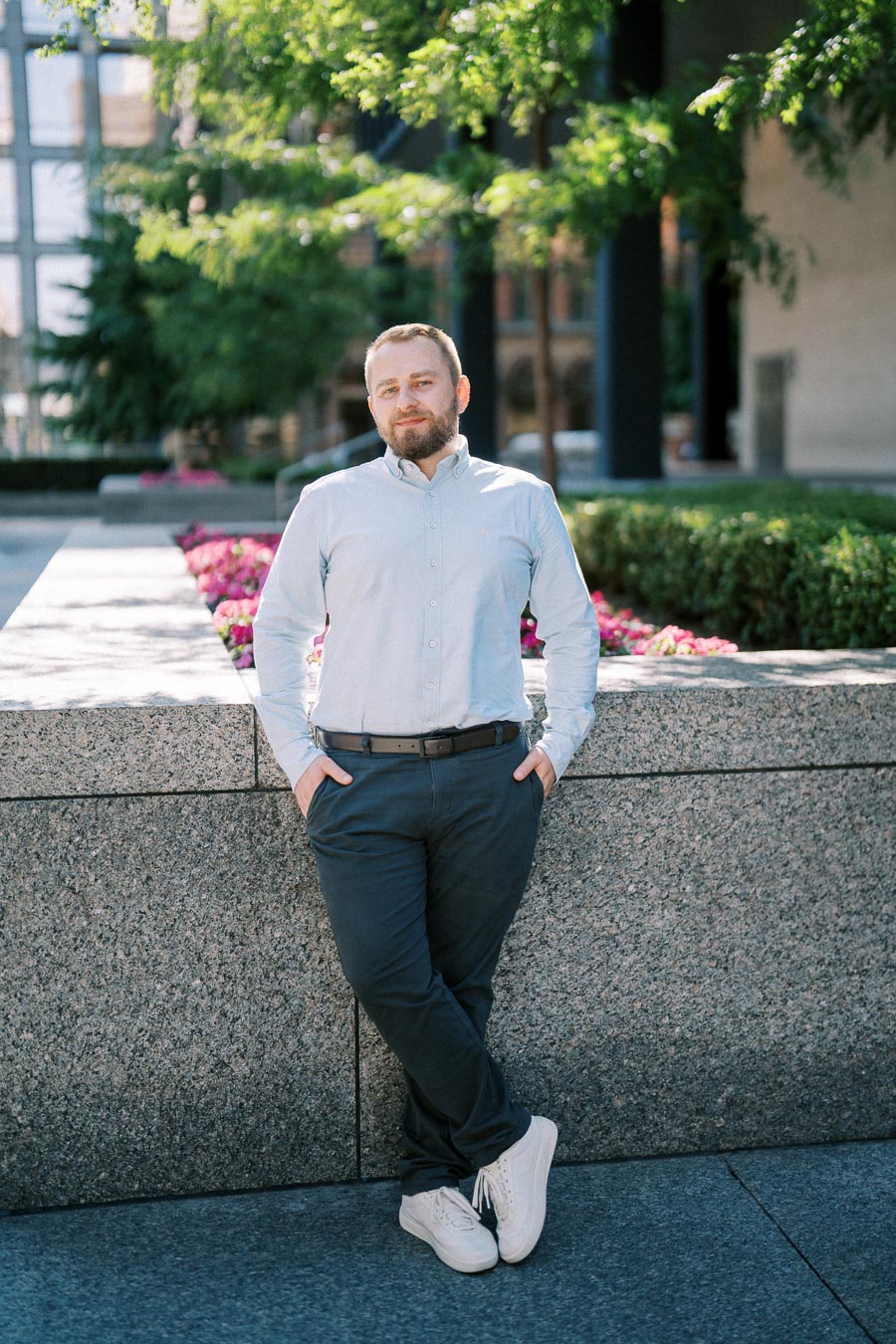 A man in a light blue shirt and dark pants stands casually against a stone wall outdoors, with vibrant pink flowers and lush greenery in the background on a sunny day.