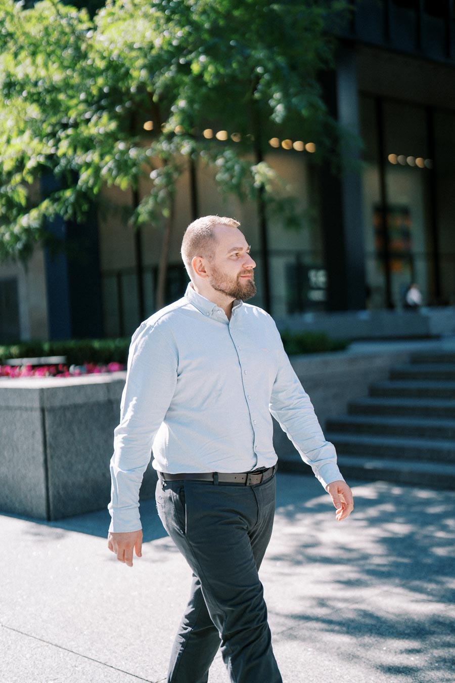 A man in a light blue shirt walks confidently outdoors on a sunny day, with office buildings and greenery in the background.