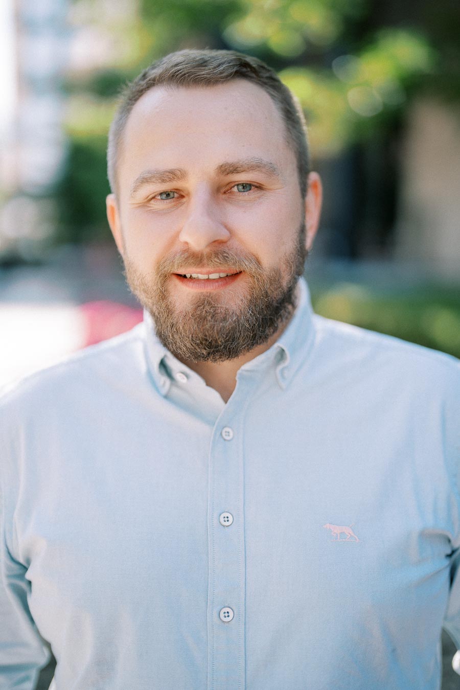 Smiling man with beard wearing light blue button-up shirt outdoors with blurred greenery in background.