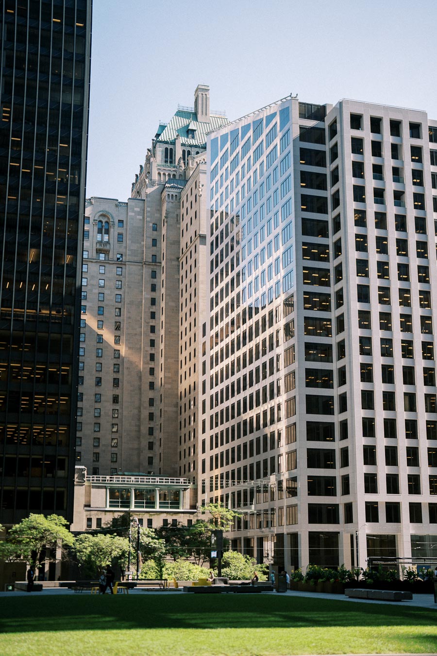 Skyscrapers and historic architecture in a vibrant urban park setting, with sunlight casting shadows on lush green lawn and trees.
