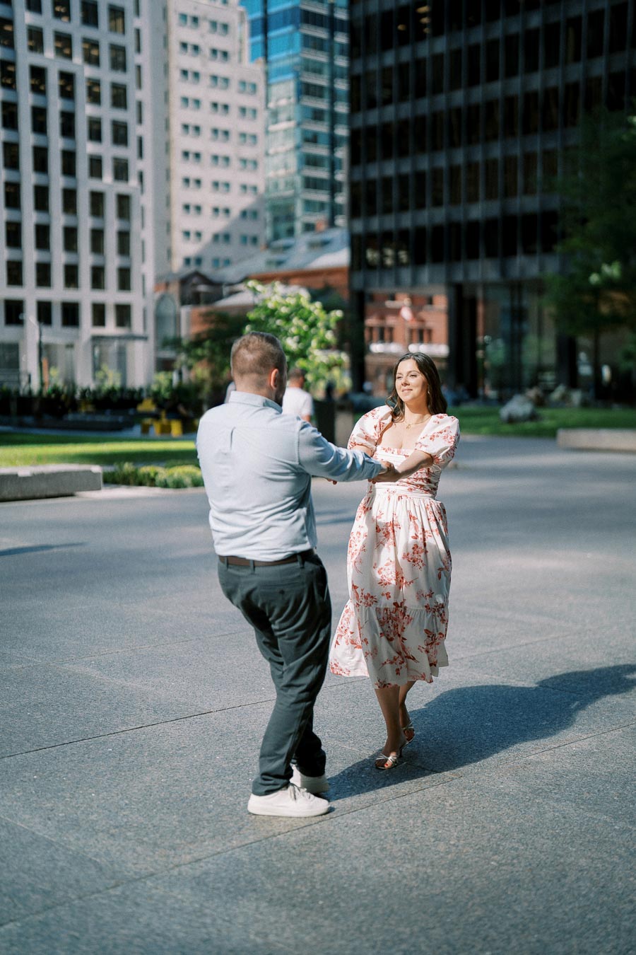 A couple dancing joyfully in an urban setting surrounded by tall buildings, with a man in a light blue shirt and dark pants spinning a woman in a floral dress, capturing a moment of happiness and connection.