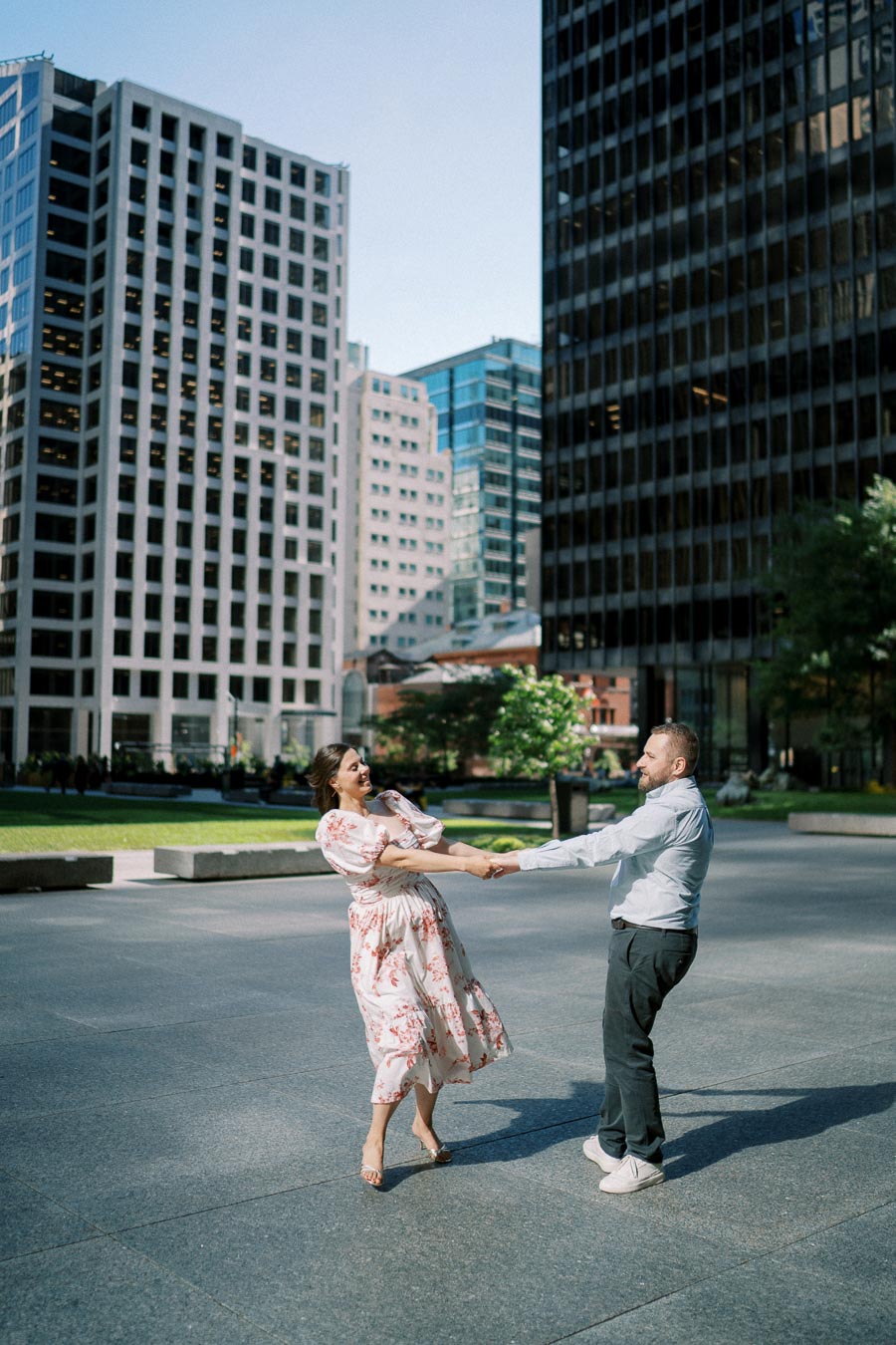 A couple joyfully dancing in an urban plaza surrounded by tall skyscrapers and greenery, enjoying a sunny day in the city.