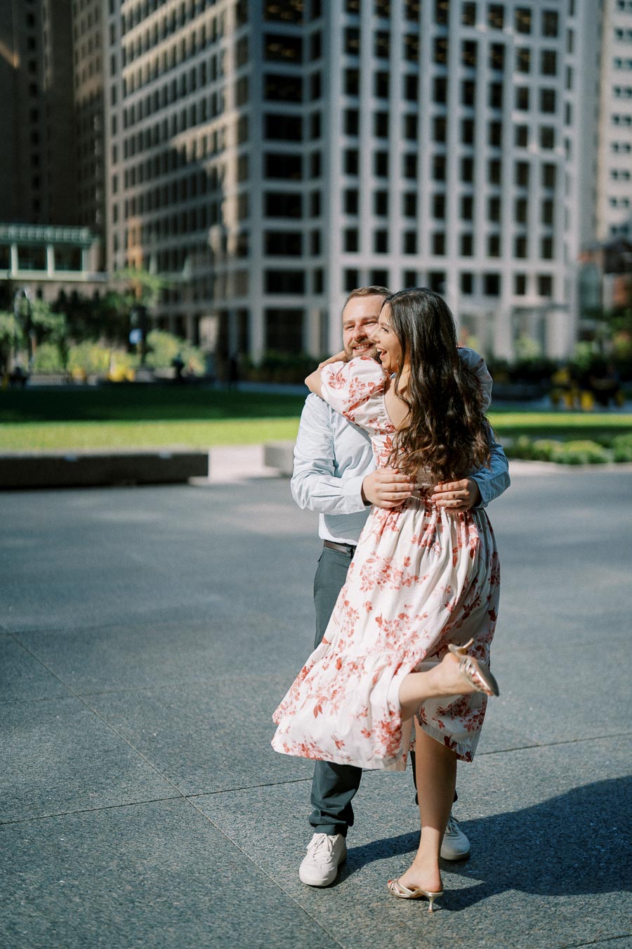A happy couple embraces and laughs in an urban setting with tall buildings in the background, surrounded by greenery and sunlight.