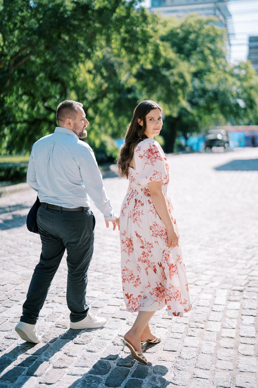 A couple walking hand in hand on a sunlit cobblestone path, featuring a woman in a floral dress and a man in a light shirt.