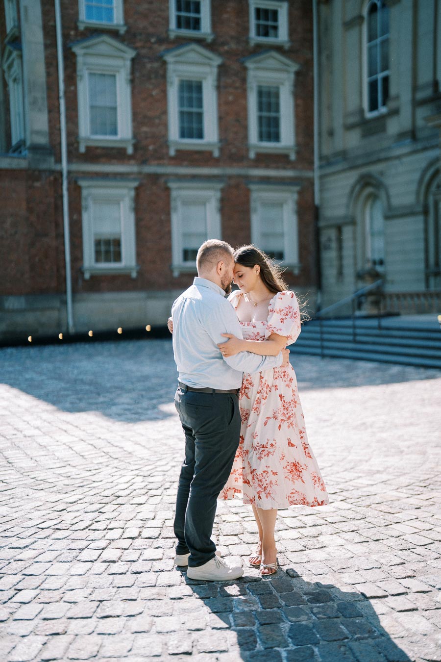 A couple embracing in a sunlit cobblestone courtyard, with a historic brick building in the background.