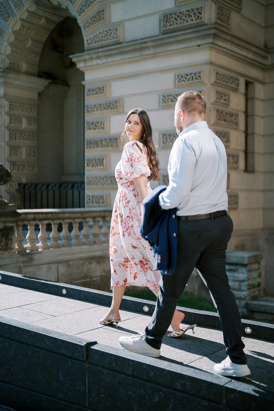 A couple walking in front of an ornate building, woman in a floral dress and man in a light blue shirt, holding a jacket.