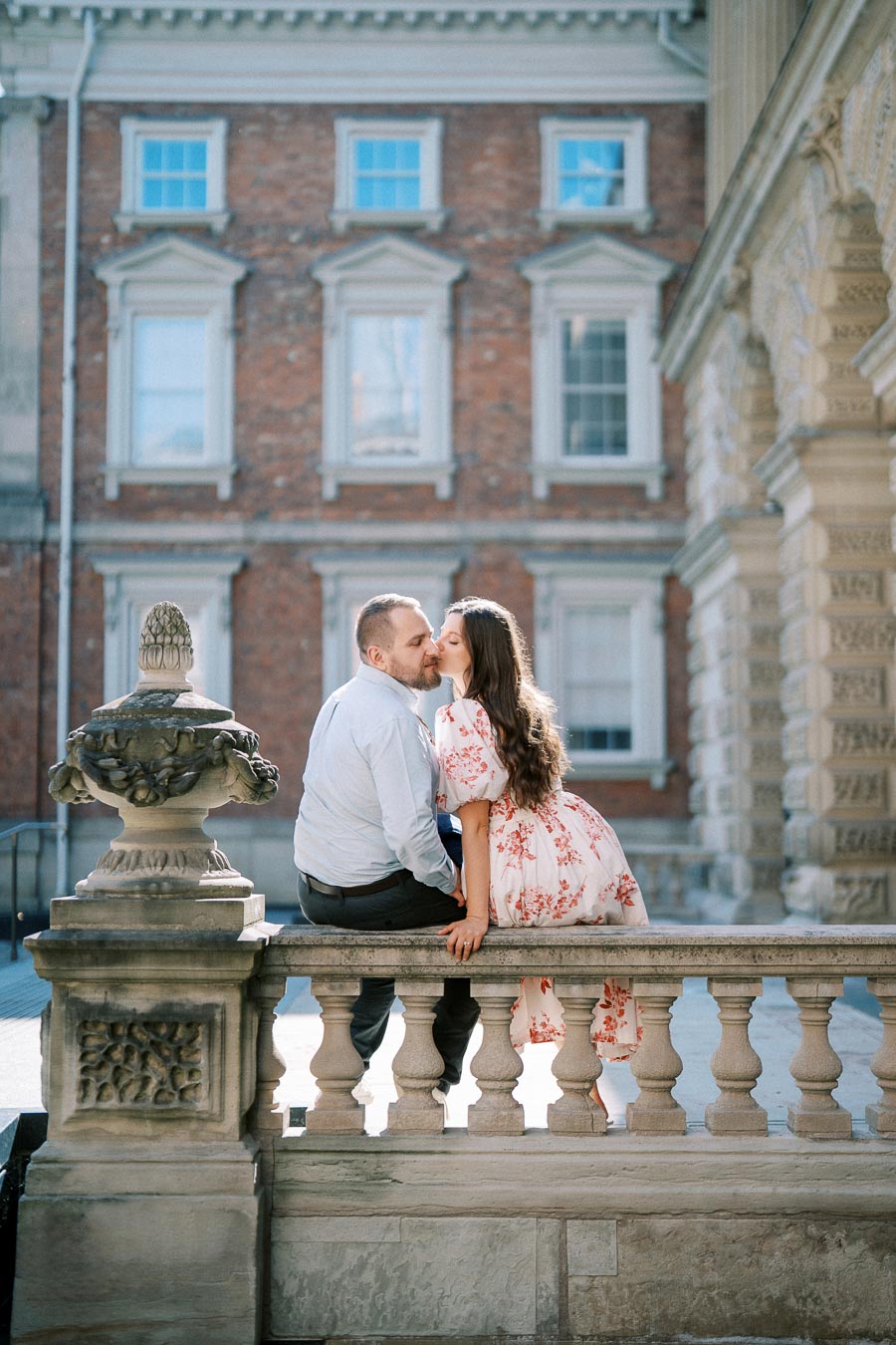 A couple shares a romantic moment on a stone balustrade in front of a historic brick building, with the woman giving the man a kiss on the cheek.
