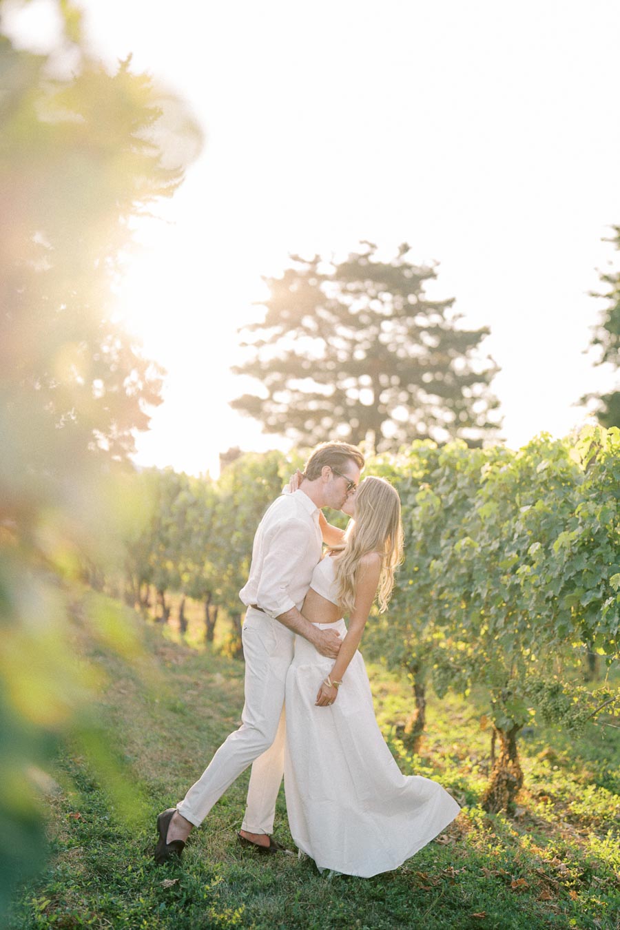 A couple in white attire shares a romantic kiss in a sunlit vineyard, surrounded by lush grapevines and trees, under a bright sky.
