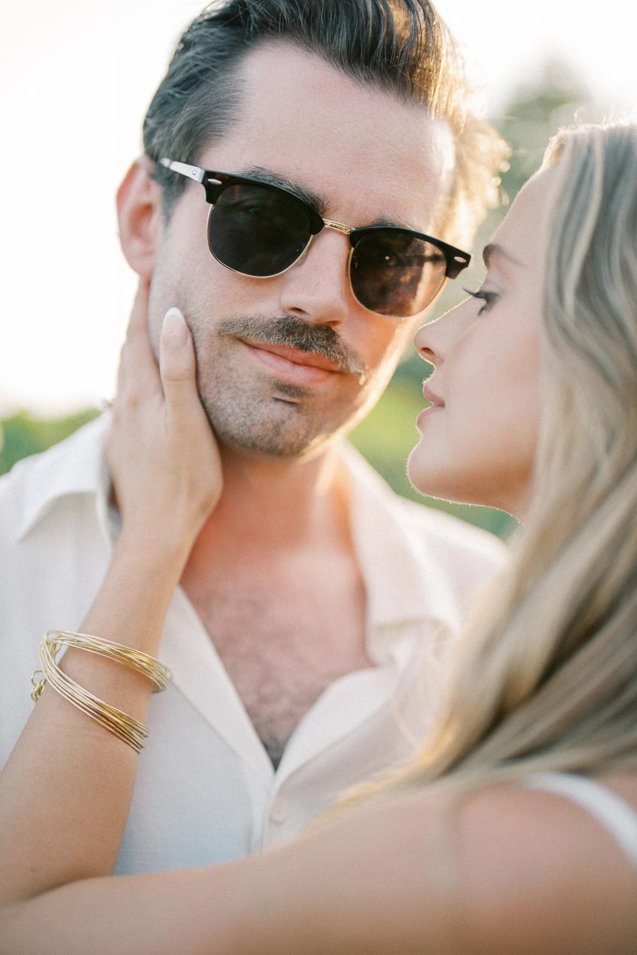Stylish couple basking in sunlight, man wearing sunglasses and white shirt with woman gently touching his face, showcasing romantic connection and summer fashion vibes.