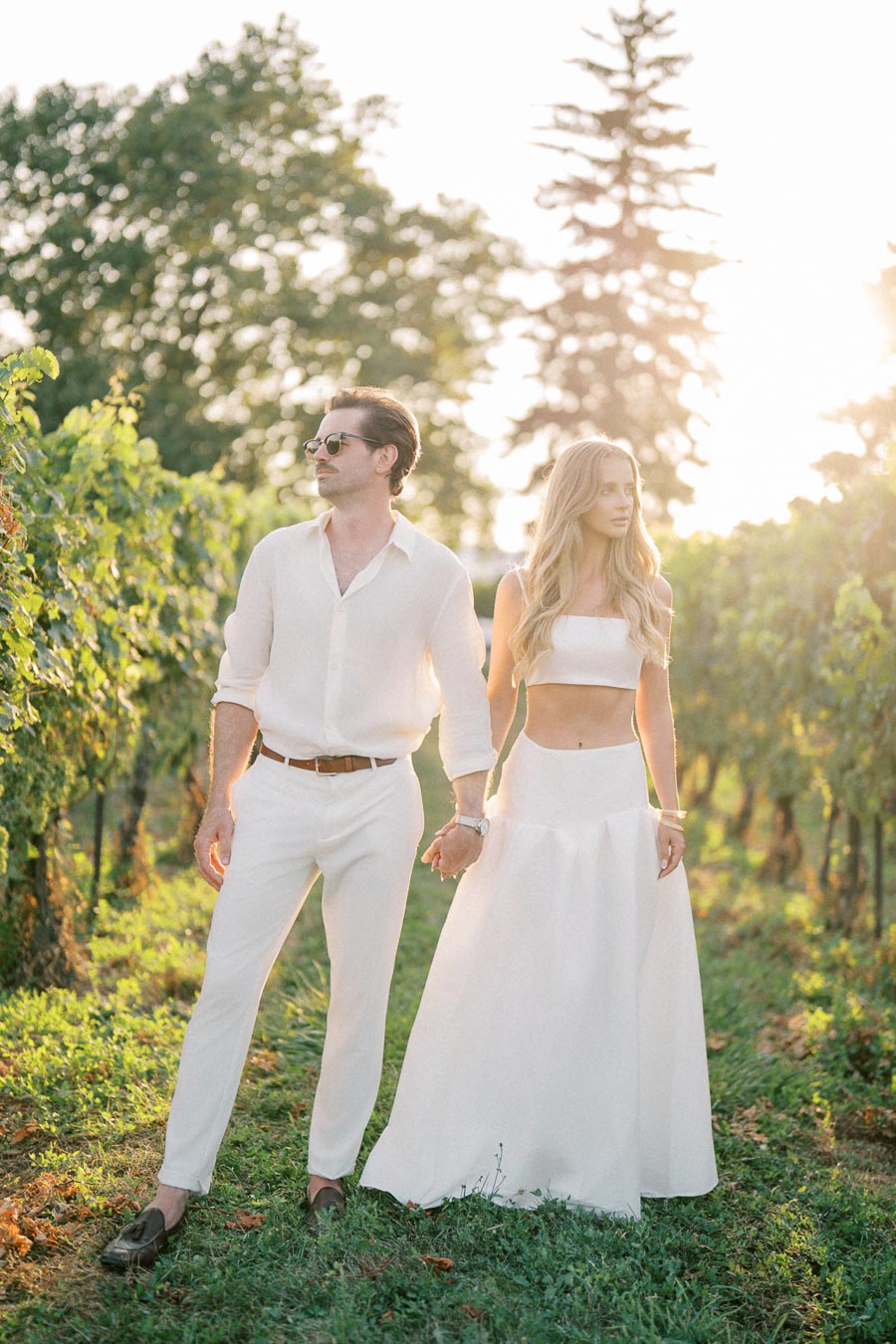 Couple in white outfits holding hands while walking through a sunlit vineyard, showcasing romance and elegance against a lush natural backdrop.