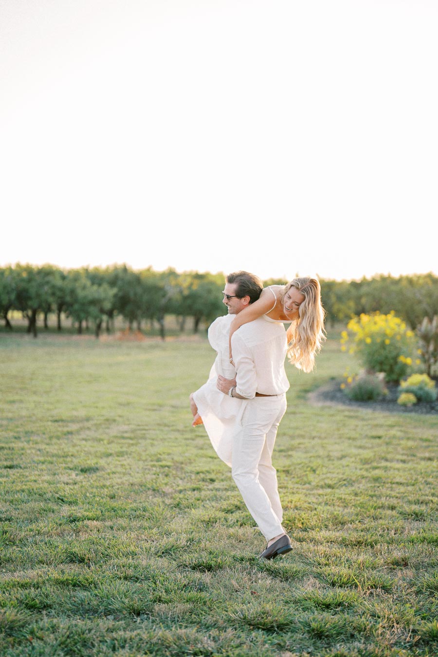 A man carrying a woman on his back in a sunny, green vineyard field, both wearing light-colored clothing and smiling joyfully.