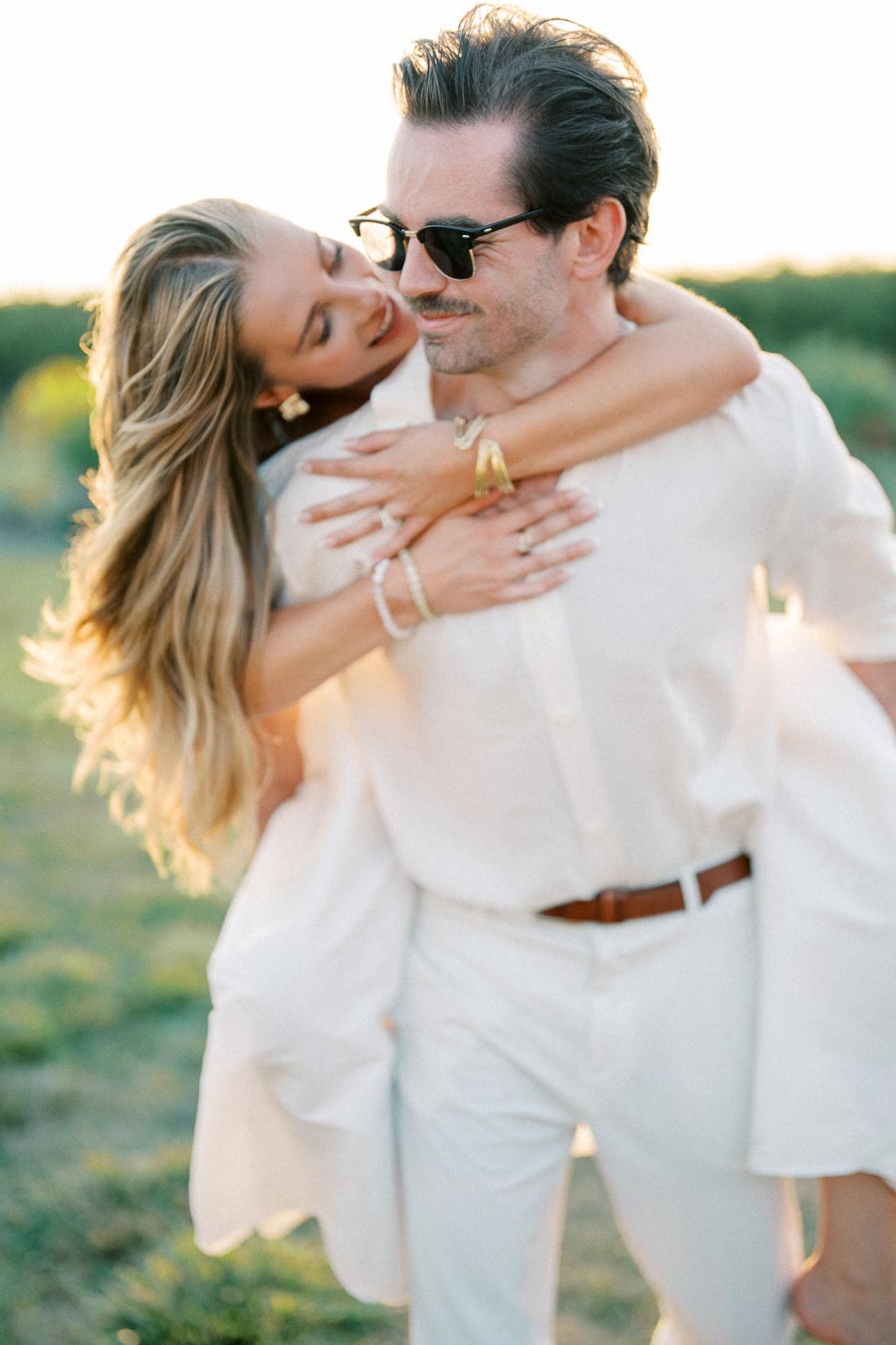 A couple enjoying a romantic moment at sunset, with the woman playfully embracing the man from behind in a sunlit field, both dressed in white.
