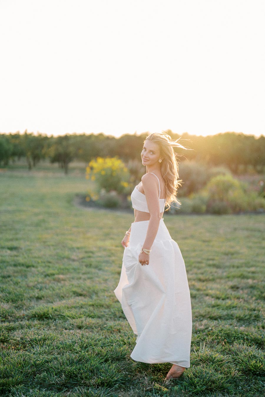 Woman in a flowing white dress standing in a sunlit meadow, looking over her shoulder with a smile at sunset.