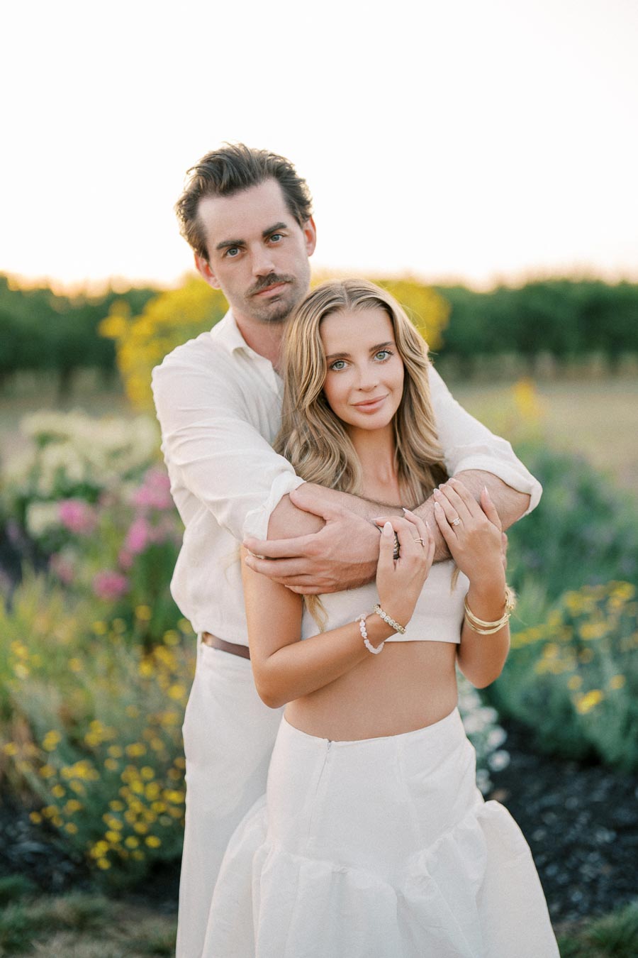 A couple embracing in a sunlit garden, wearing coordinated white outfits, surrounded by blooming flowers and greenery.