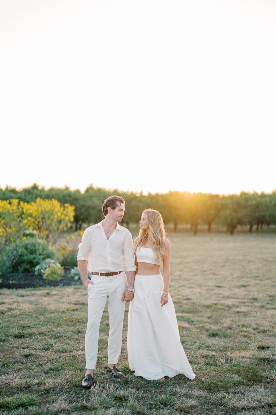 Couple in white outfits walking hand-in-hand through a sunlit field, surrounded by greenery and basking in a romantic sunset.