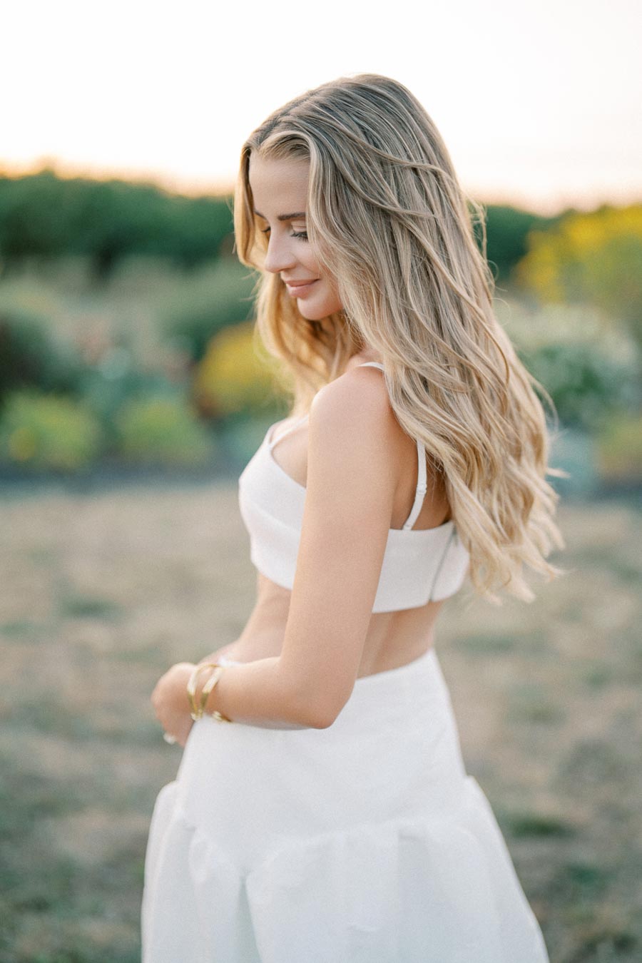Smiling woman with long blonde hair in a white outfit looking down in an outdoor setting during golden hour