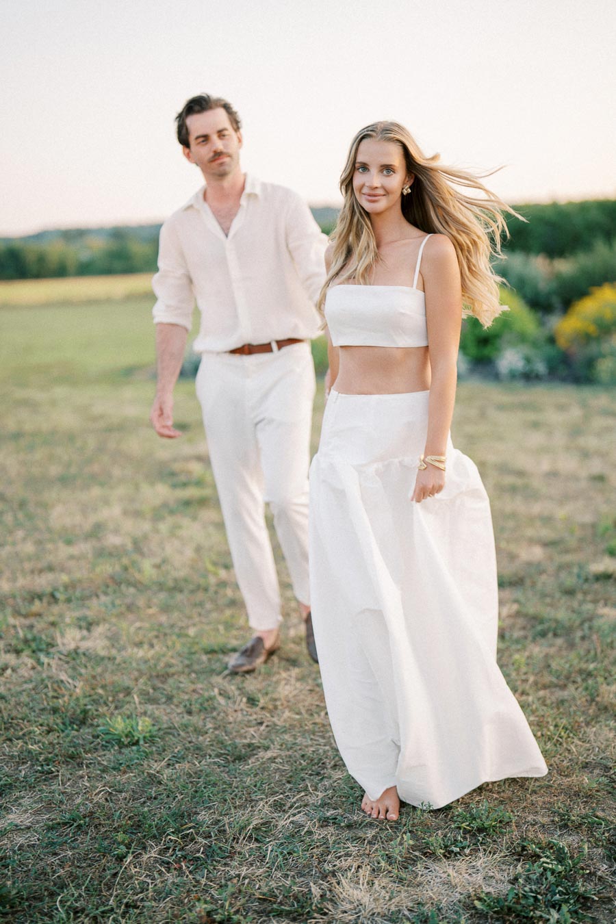 A couple in matching white outfits walking through a grassy field at sunset, creating a romantic and serene atmosphere.