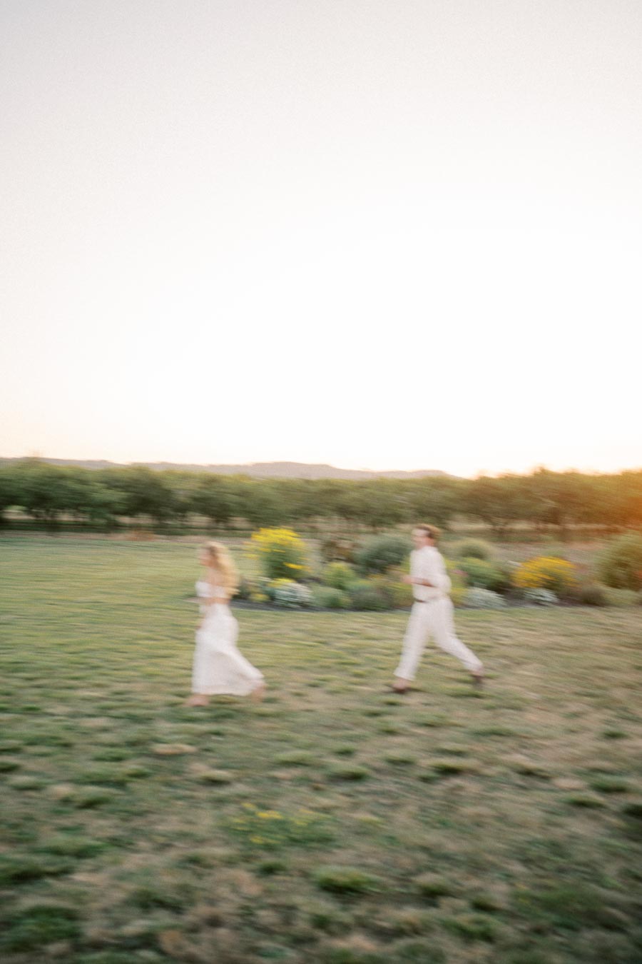 Blurred image of two people in white attire running across a grassy field with vibrant flora, set against a backdrop of a hazy sunset.
