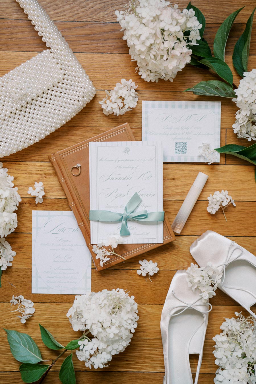 Flat lay of elegant wedding invitations tied with a blue ribbon, a pearl handbag, white high heels, and delicate white hydrangea flowers on a wooden floor for a stylish bridal theme.