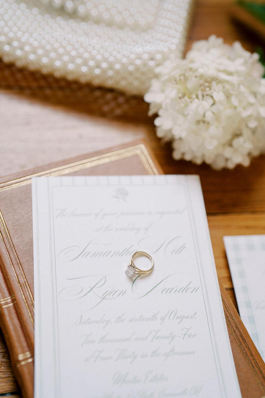 A close-up of an elegant wedding invitation with cursive text, featuring a gold engagement ring resting on top, surrounded by soft-focus details including a white floral arrangement and books, all on a wooden surface.