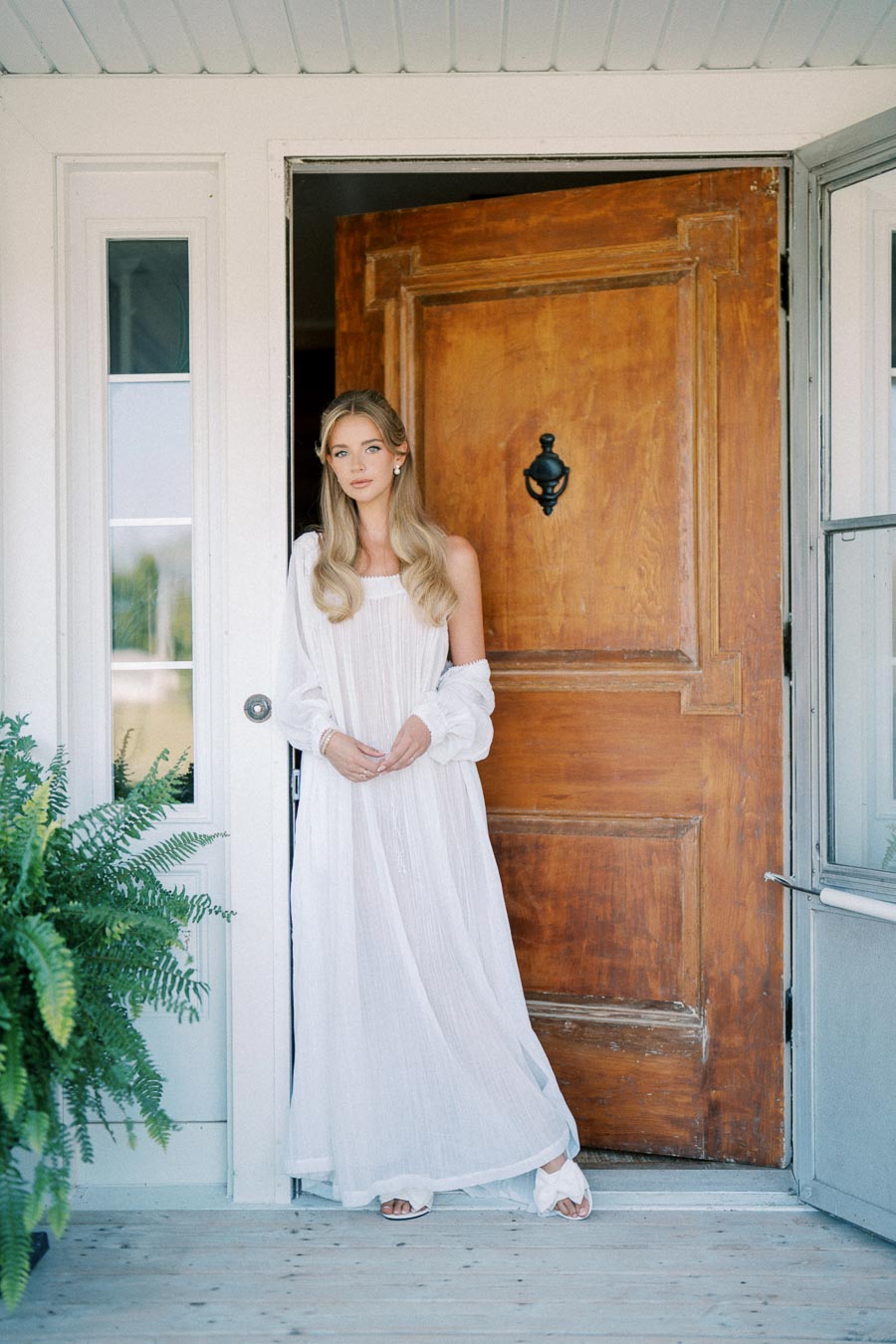 A woman in a long white dress stands in a wooden doorway, exuding an ethereal, serene vibe. The entrance is accentuated by a large, rustic door, flanked by glass panels and lush green plants, contributing to a classic, elegant look.