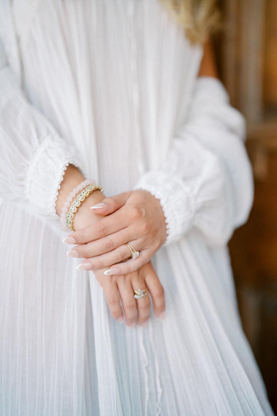 Close-up of a woman's hands adorned with elegant gold rings and a stylish bracelet, wearing a flowing white dress with lace details.