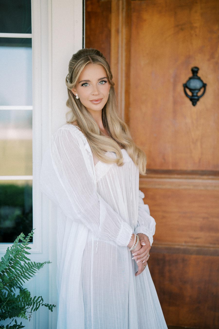 Elegant woman in a white flowing dress standing by a wooden door with a fern plant, showcasing soft vintage styling and natural beauty.