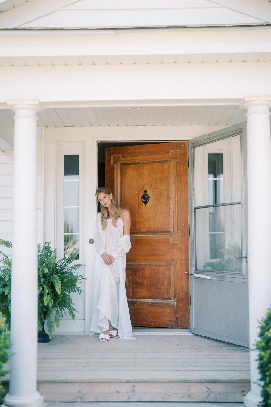 Young woman in a flowing white dress standing on a porch with wooden front door, surrounded by white pillars and lush greenery.