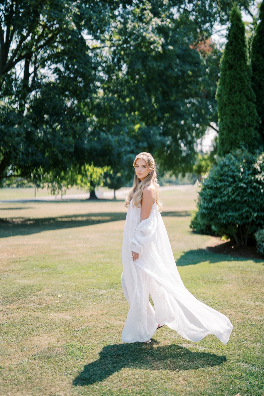 A woman in a flowing white dress walks gracefully on a lush green lawn, surrounded by tall trees and vibrant bushes on a sunny day.