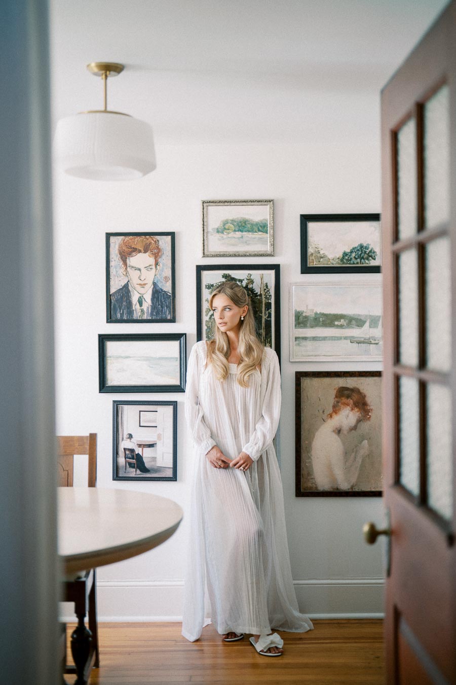A woman in a flowing white dress stands in a bright, modern room surrounded by a gallery wall of framed artwork, exuding a serene and artistic ambiance.