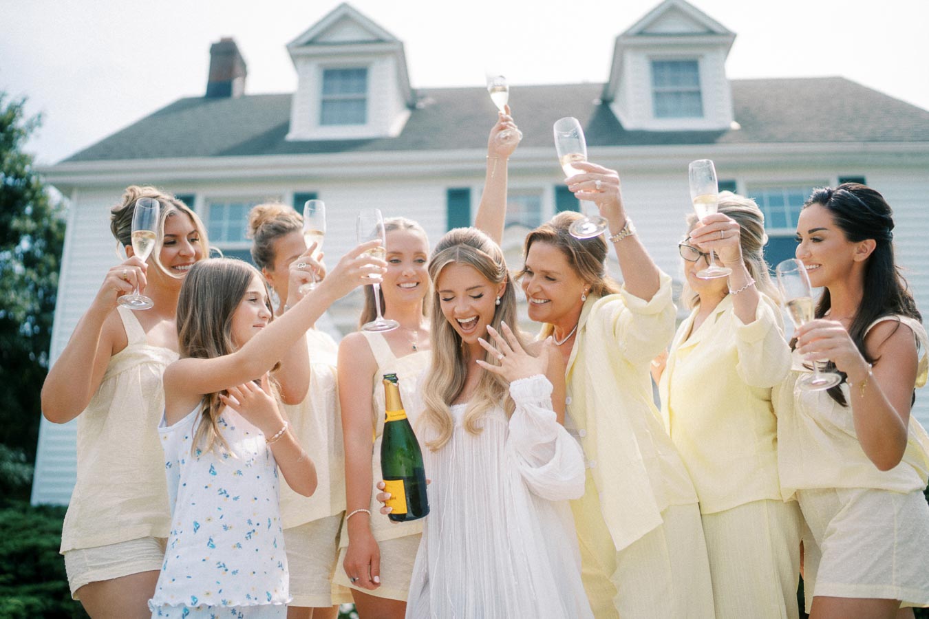 A joyful group of women celebrating an engagement outdoors, with one woman showing off her ring and holding a champagne bottle, while others raise their glasses in cheers, all dressed in light-colored outfits in front of a charming house.