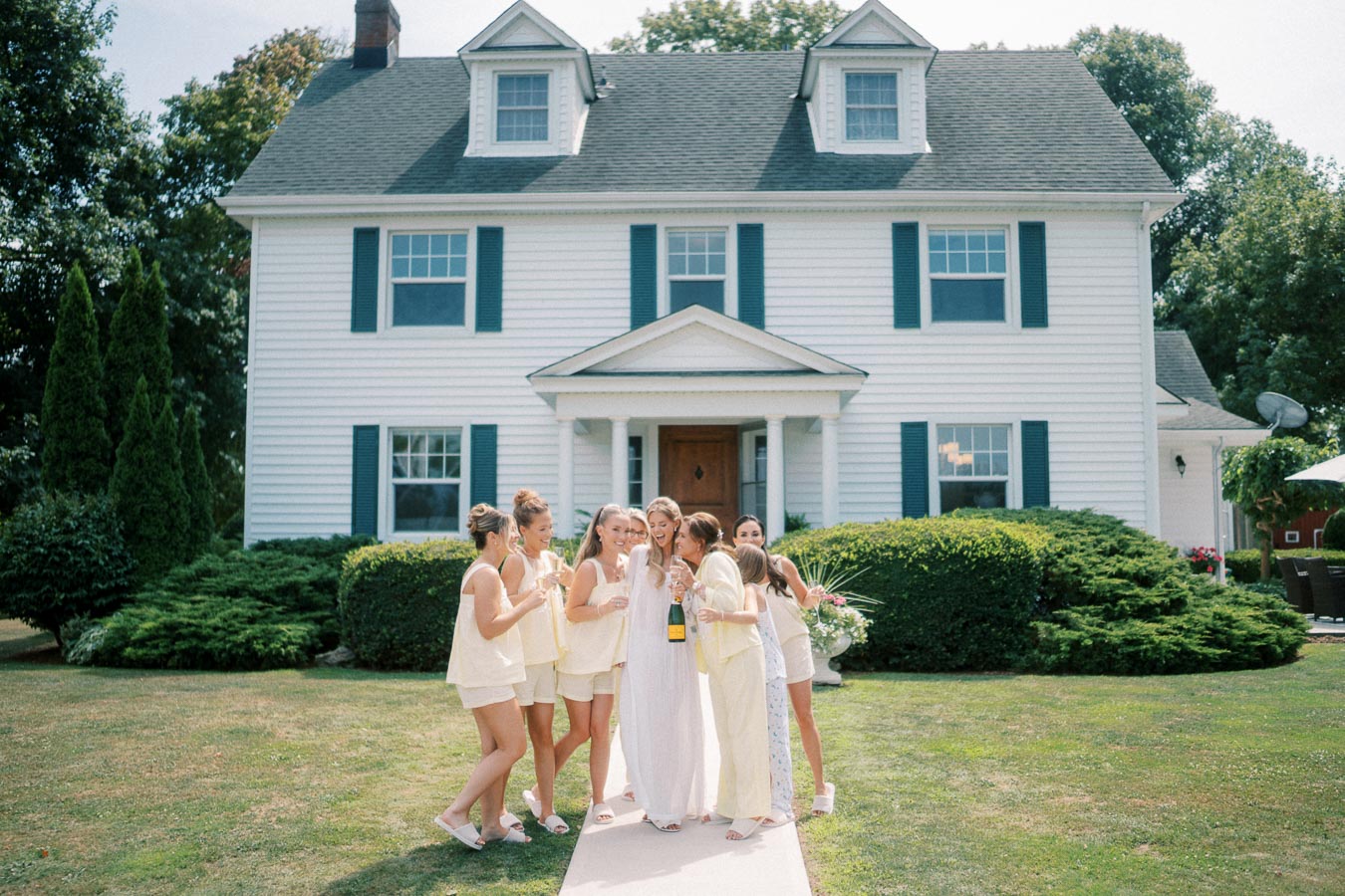 Bridal party in matching outfits celebrating with champagne in front of a large white colonial-style house.