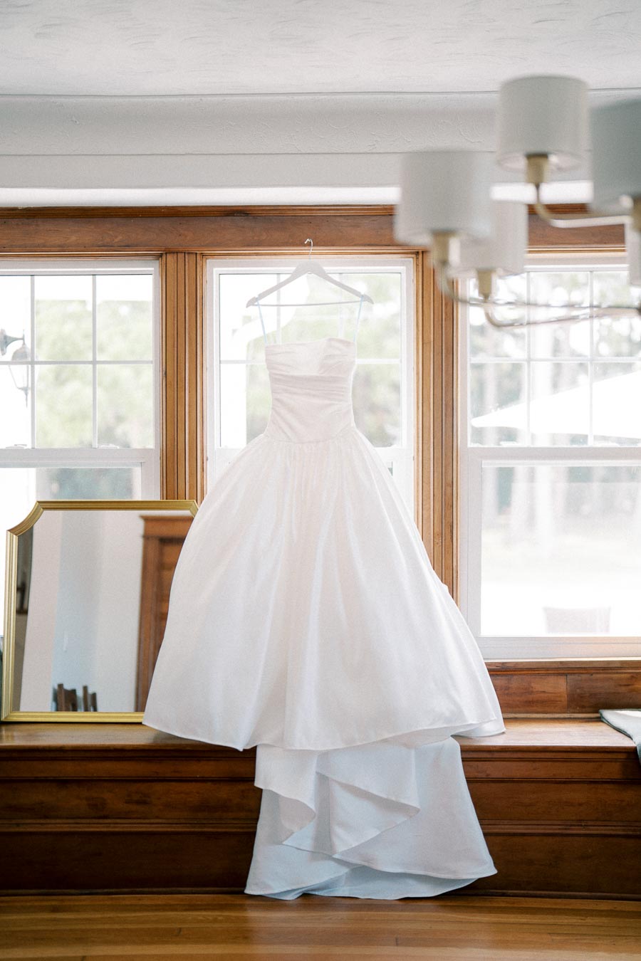 Elegant white wedding dress hanging on a window frame, illuminated by natural light, with a full skirt and strapless bodice, ideal for bridal fashion inspiration.
