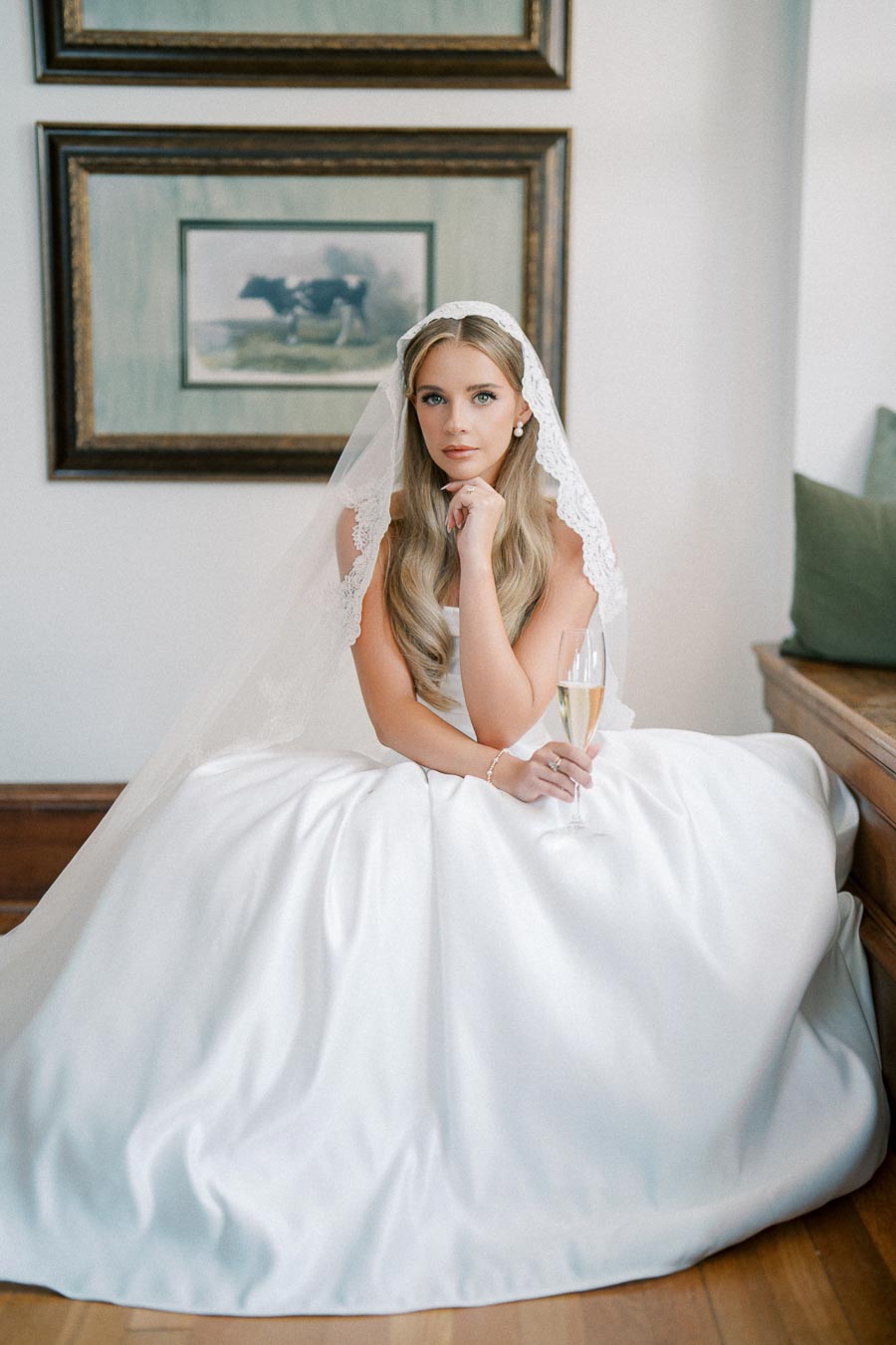 Bridal portrait of a woman in an elegant white wedding dress and lace veil, holding a champagne glass, seated indoors against framed wall art.