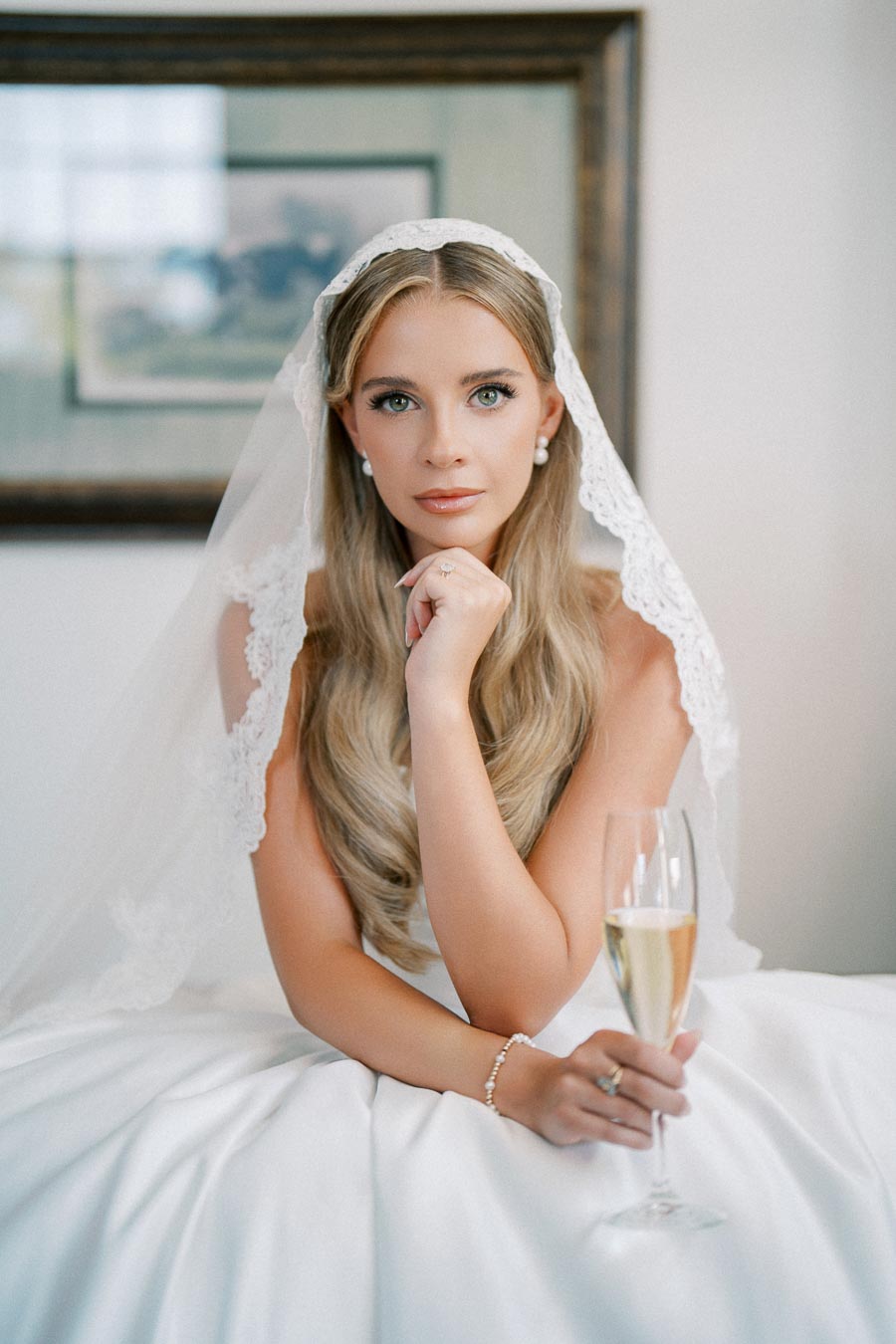 Bridal portrait of a woman seated, wearing a white lace veil and gown, holding a champagne glass, with long blonde hair and pearl earrings.