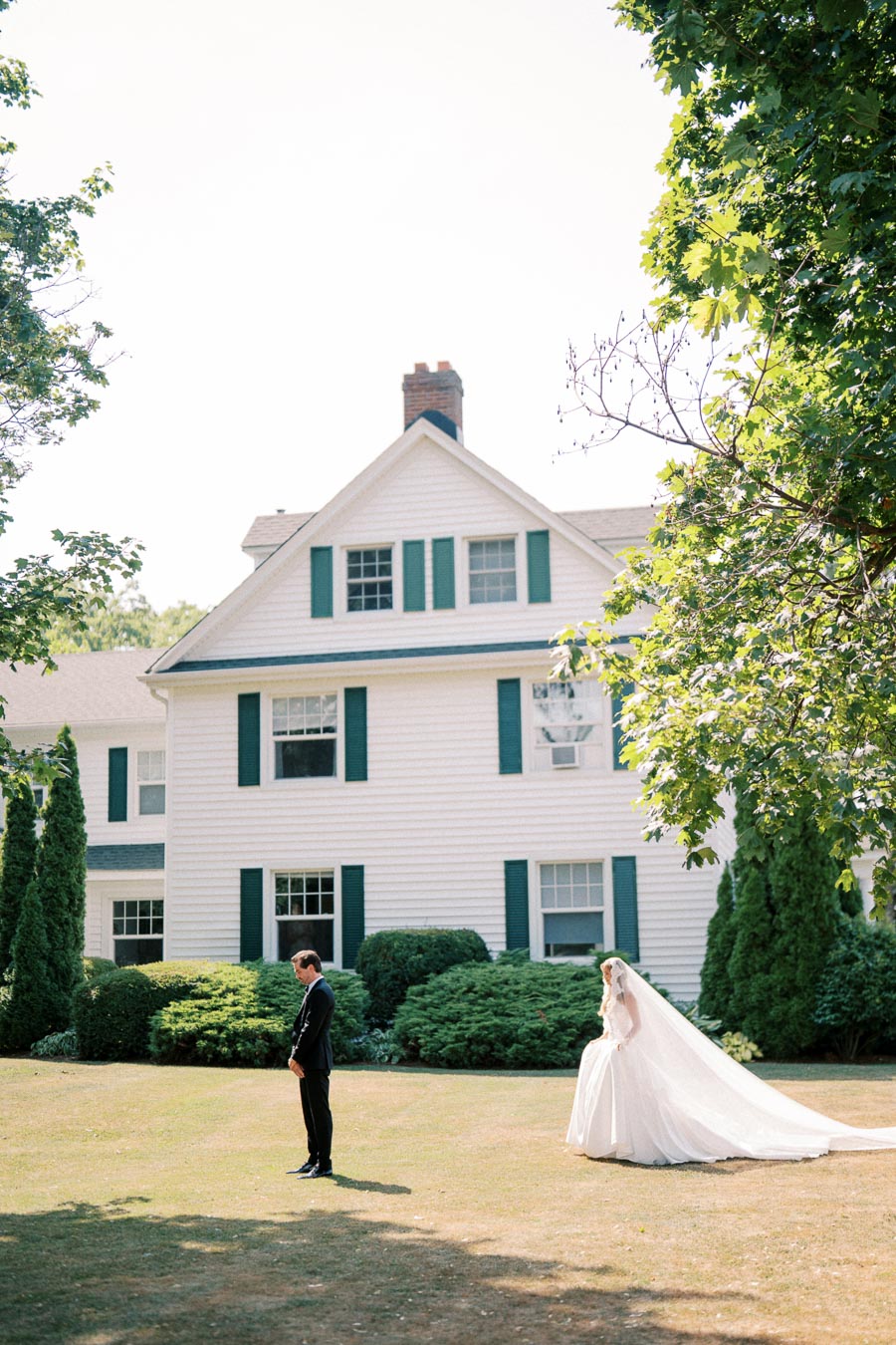 A bride in a flowing white gown and veil approaches a groom in a black suit during a wedding day first look, set against the backdrop of a classic white house with green shutters surrounded by lush greenery and trees.
