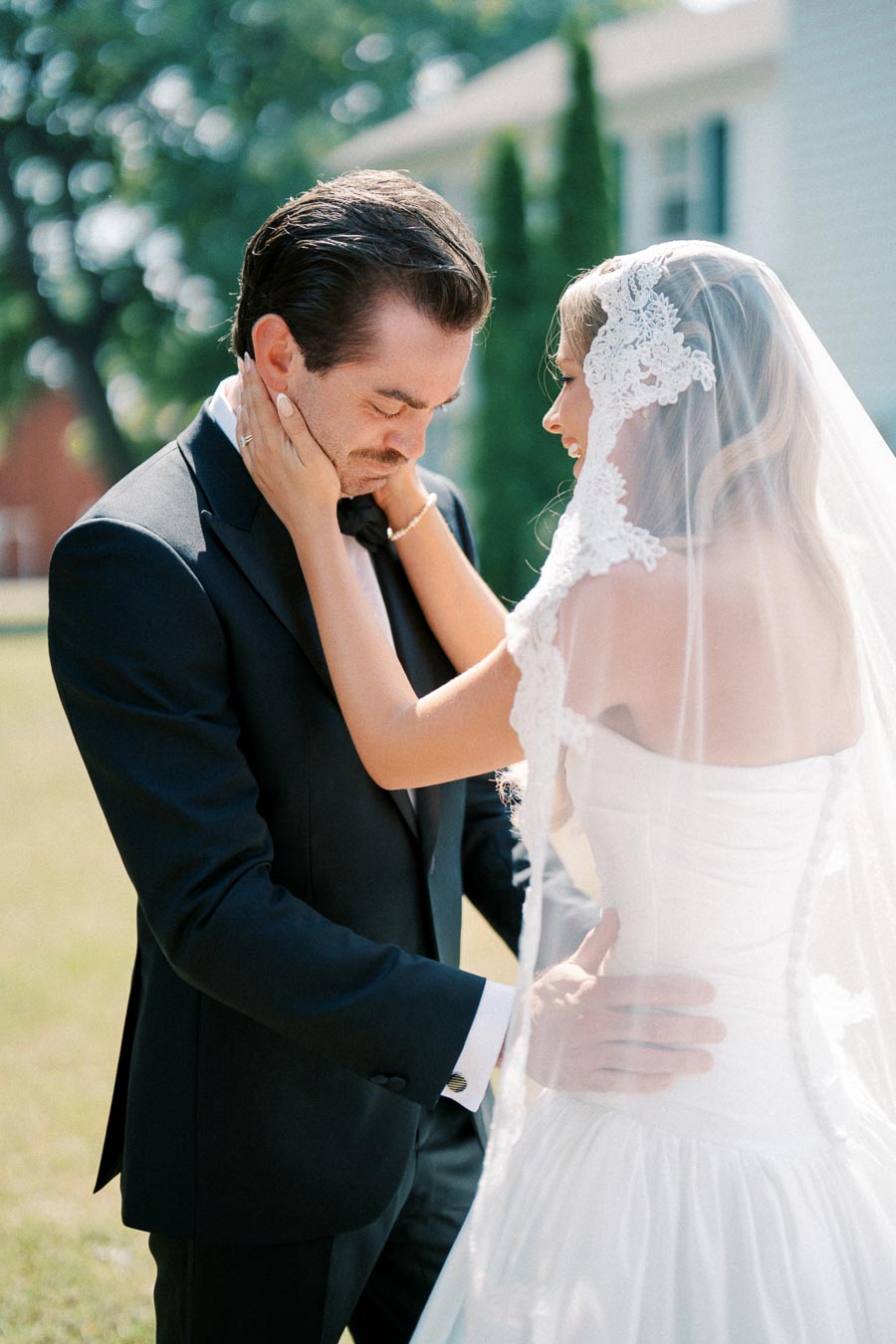 Bride and groom sharing a tender moment outdoors on their wedding day, with the bride in a lace wedding dress and veil, and the groom in a black tuxedo.