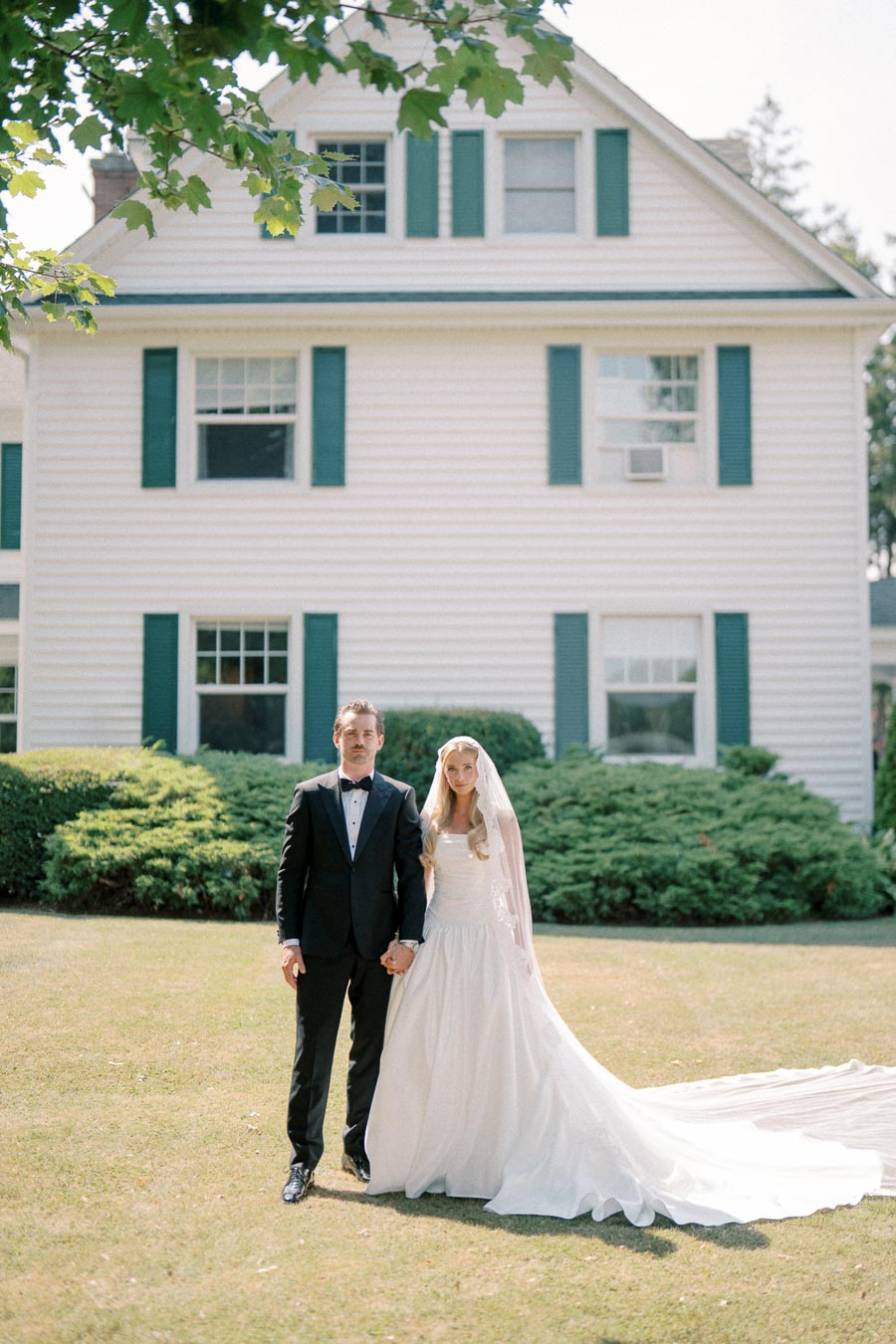 Elegant wedding portrait of a bride in a flowing white gown and veil and groom in a black tuxedo standing in front of a large white house with green shutters, surrounded by lush greenery and a manicured lawn.
