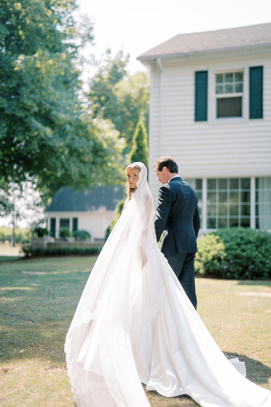 A bride in a flowing white gown and veil walks with a groom in a dark suit in a garden setting, with a charming white house in the background.