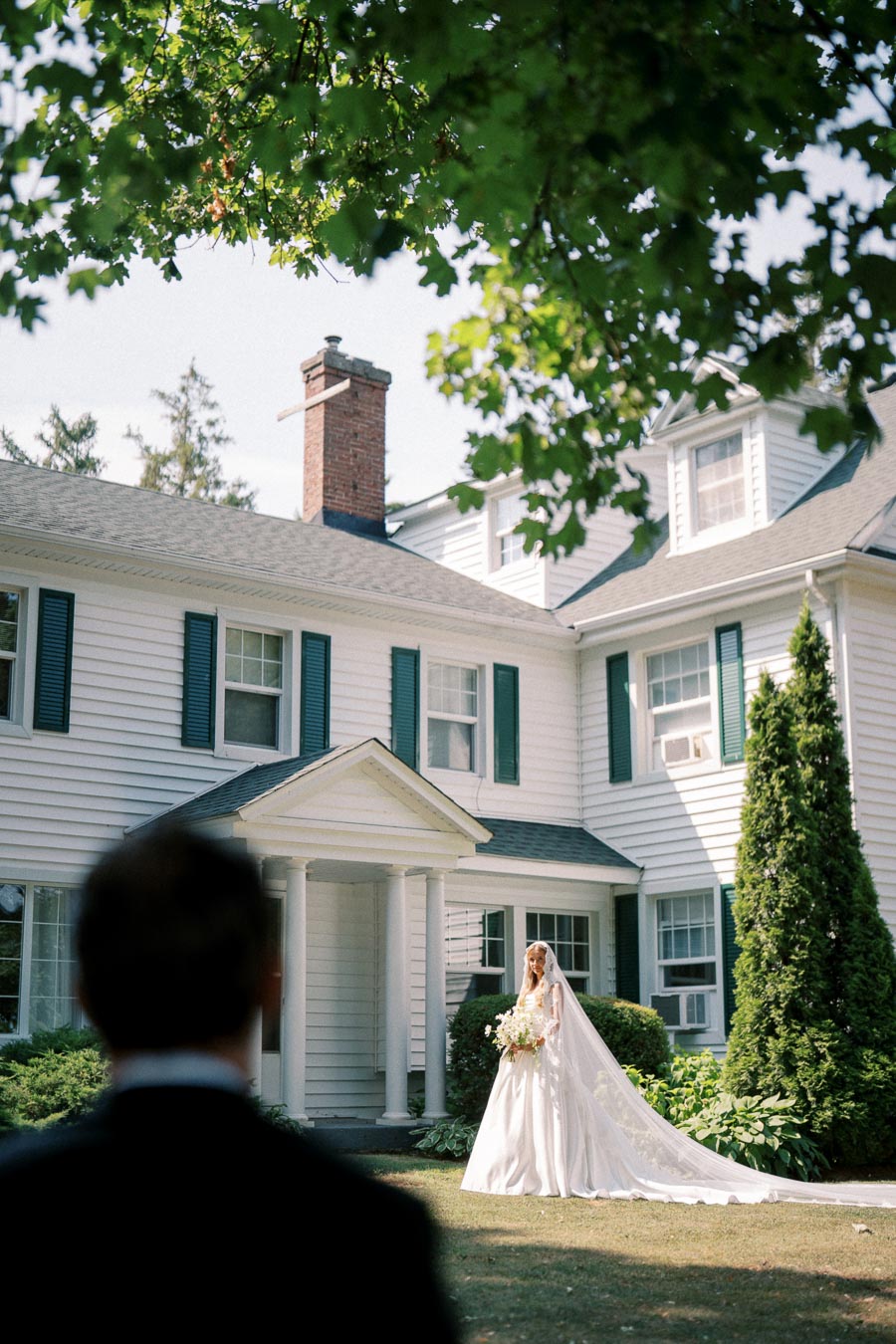 Bride in elegant white gown standing in front of a classic white house with green shutters, surrounded by lush greenery, under a partially sunny sky.