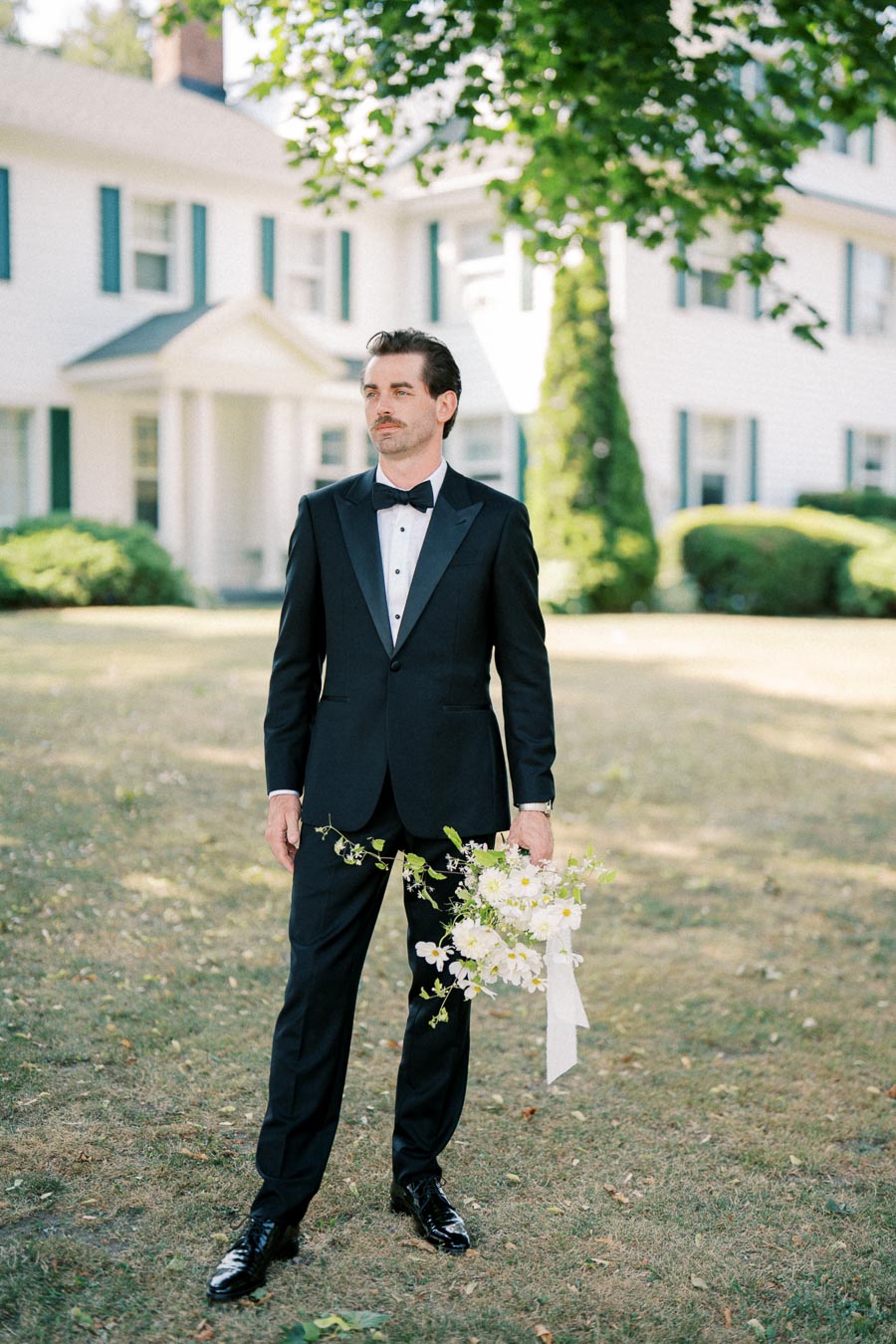A man in a formal black suit holding a bouquet of flowers stands outside in front of a large white house with green shutters, surrounded by greenery.