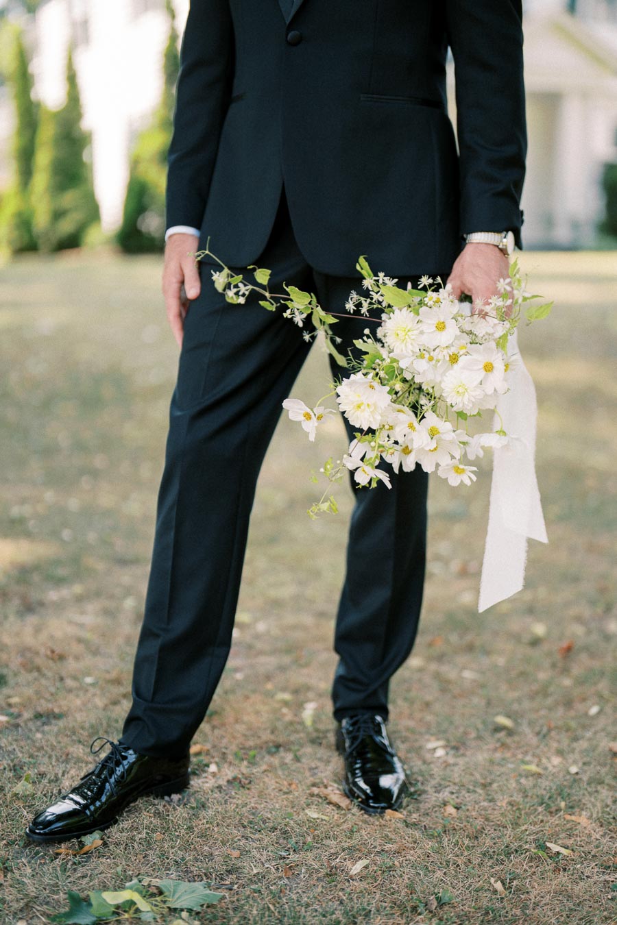 A man in a black suit holds a bouquet of white flowers with green leaves and ribbon, standing on grass in an outdoor setting.