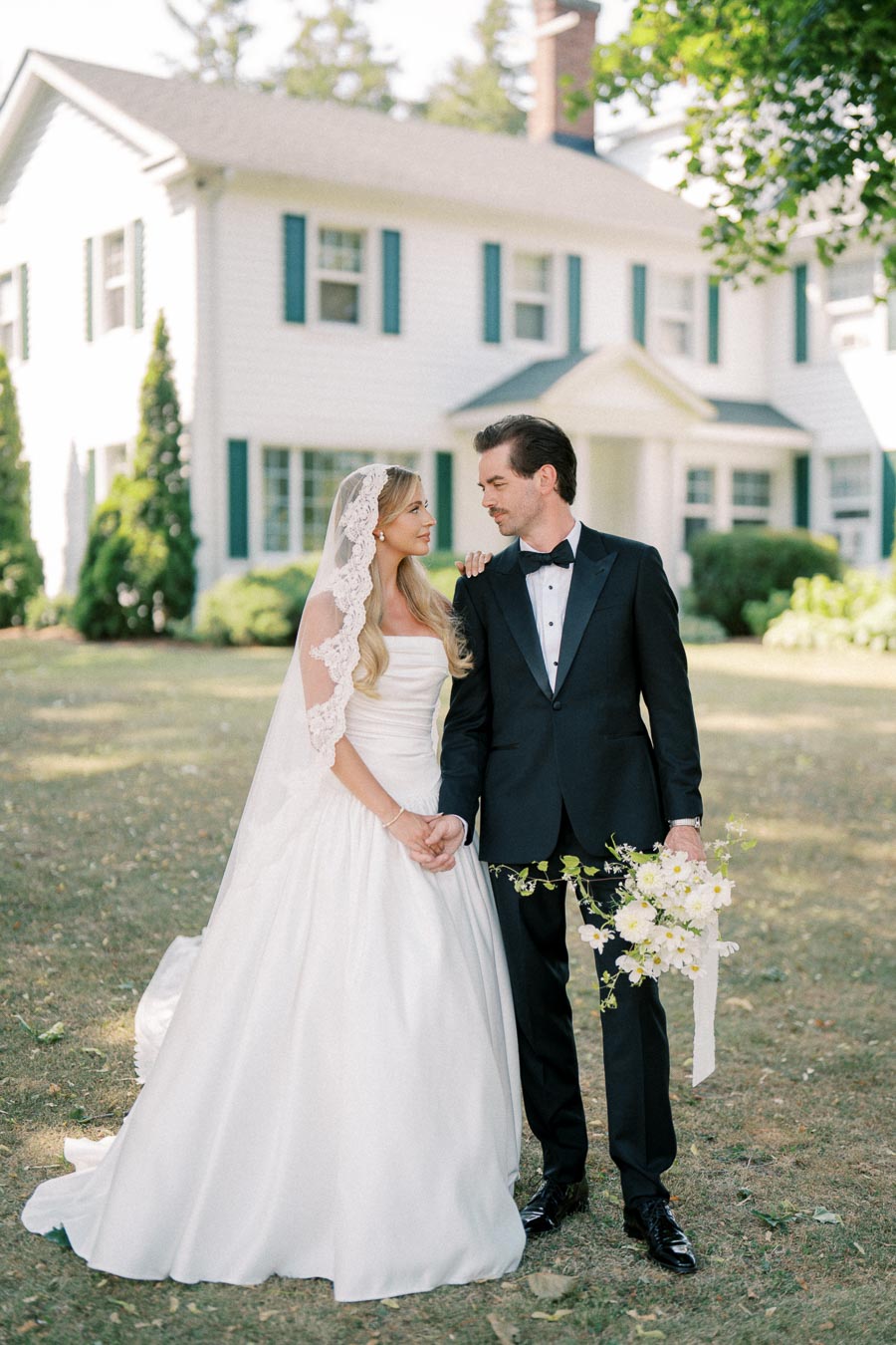 Elegant bride and groom in classic wedding attire standing in front of a traditional white house, surrounded by lush greenery, capturing a timeless romantic moment.