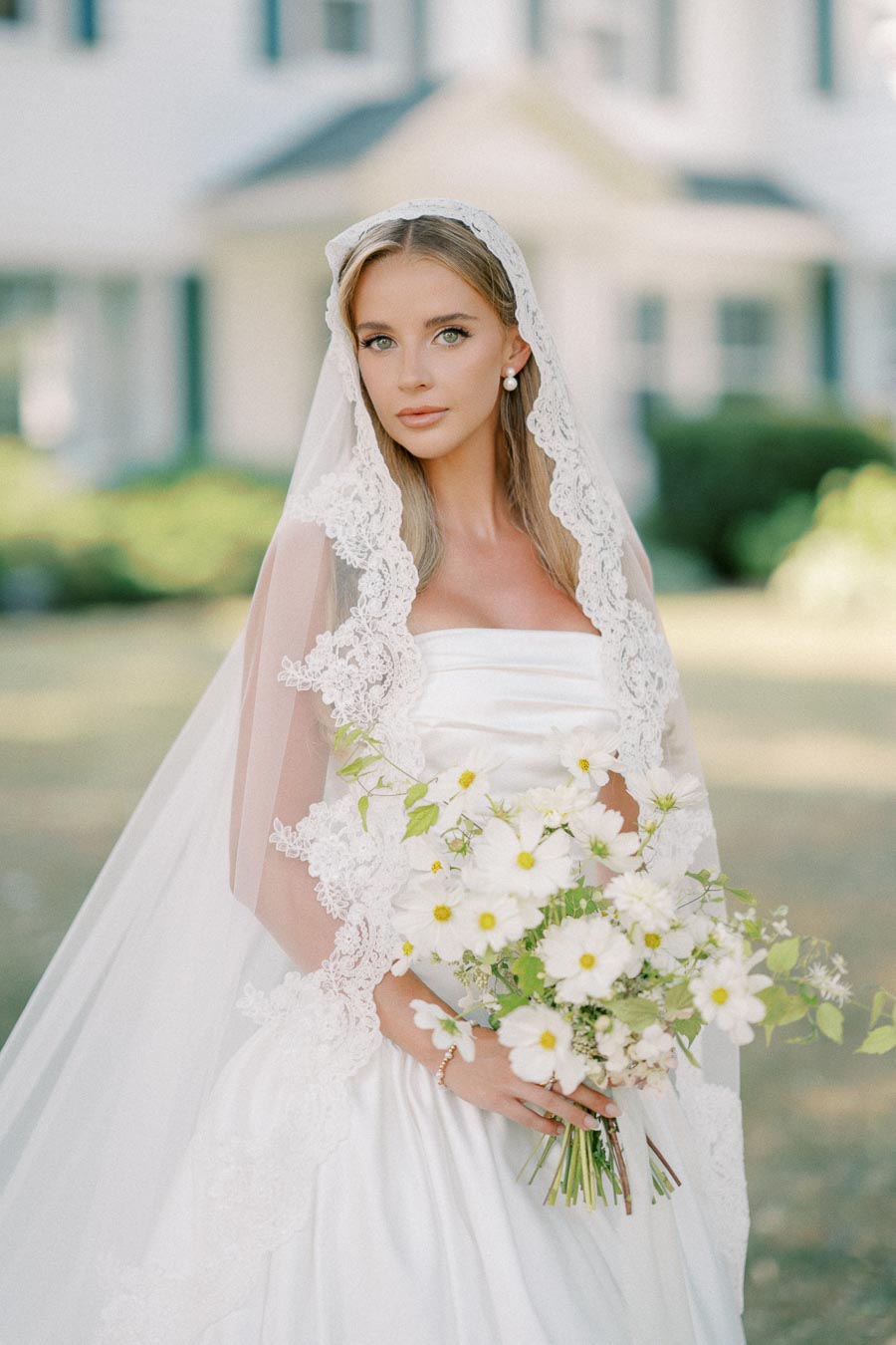 Bride in an elegant white wedding dress and lace veil holding a bouquet of white flowers in an outdoor setting.
