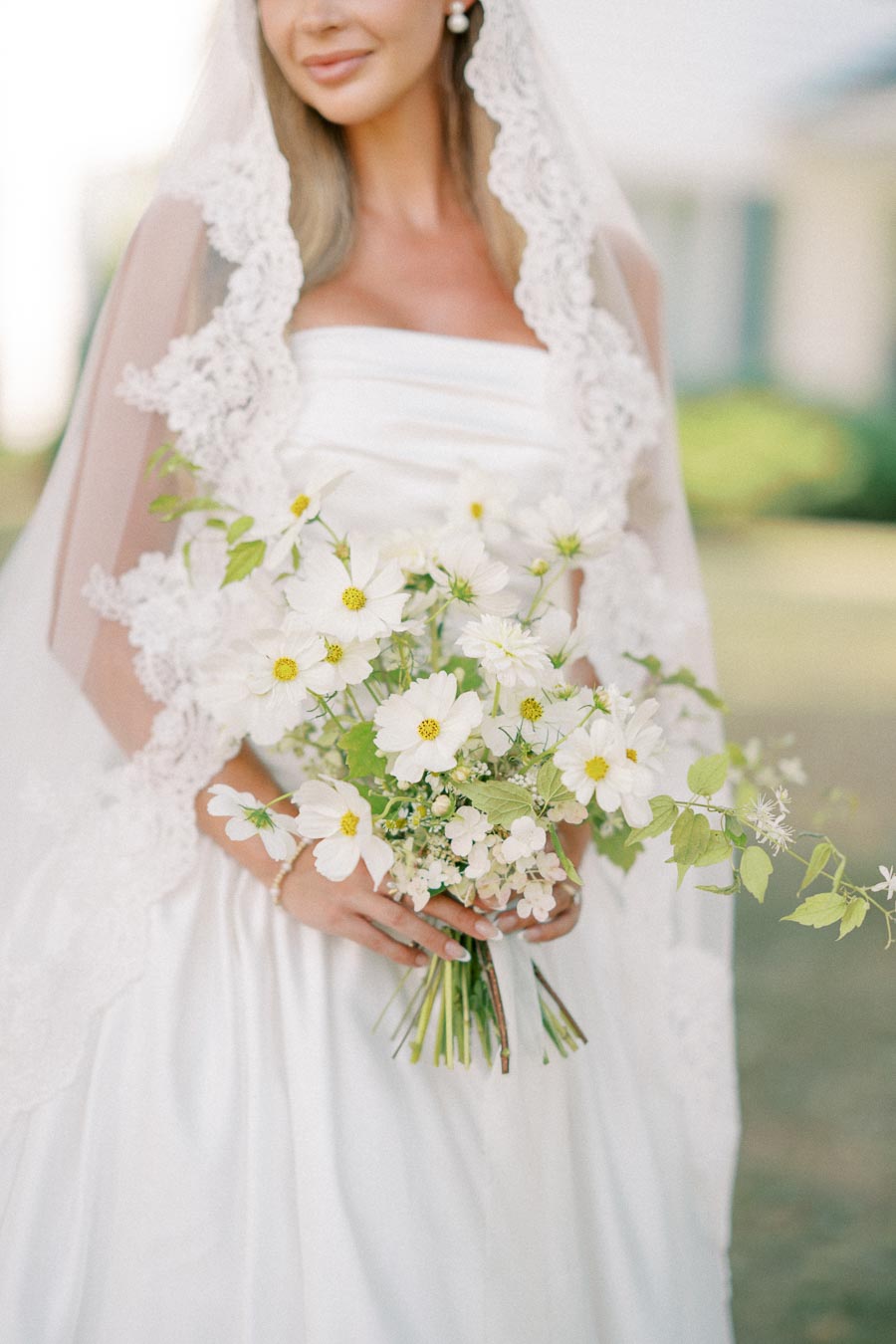 Bride holding a bouquet of white flowers, dressed in a detailed lace veil and elegant gown.