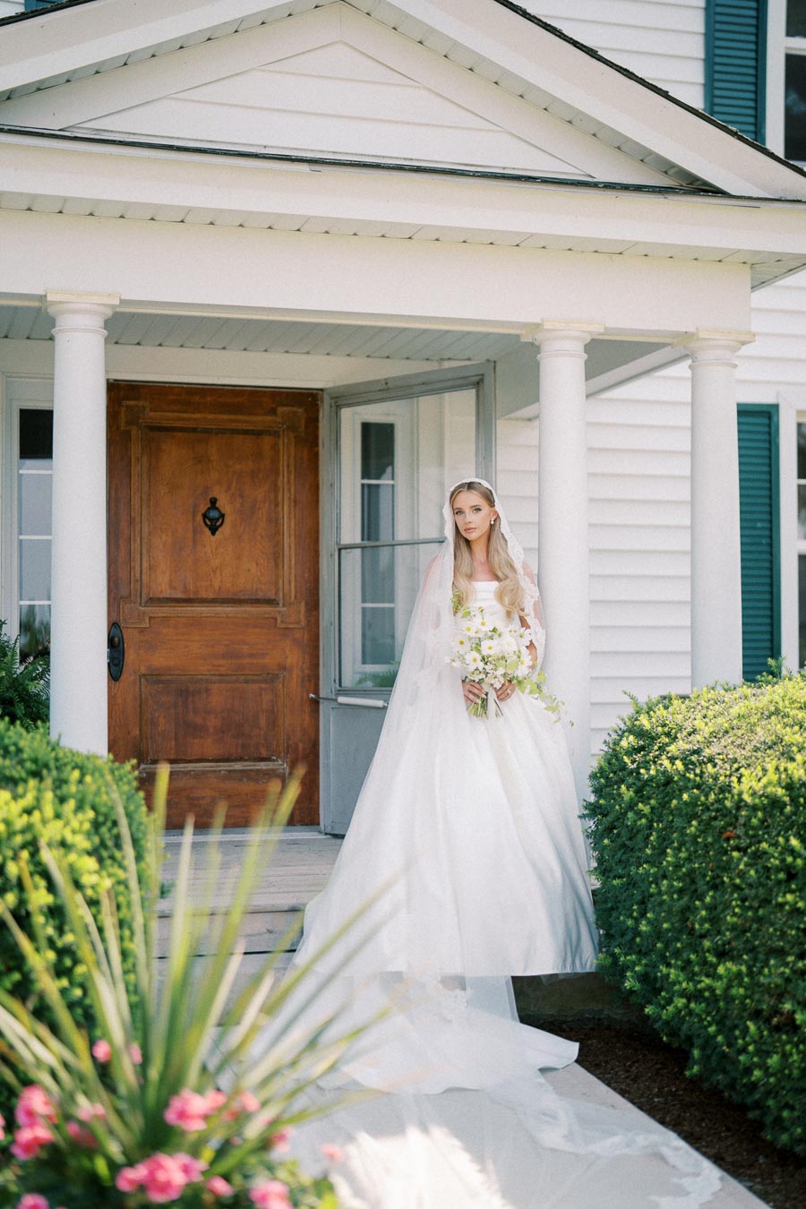 Bride in elegant white gown holding a bouquet, standing in front of a charming house with wooden door and white pillars, surrounded by lush greenery and flowers.